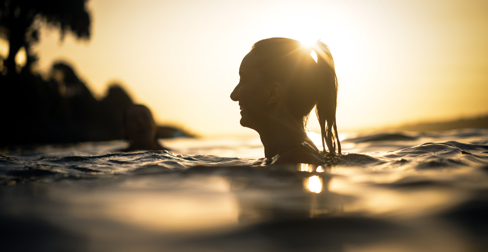 A scenic coastal view featuring a wide expanse of calm blue water under a clear sky. In the foreground, rugged rocky cliffs extend into the sea, with patches of green vegetation scattered across them. The horizon line is visible in the distance, where the ocean meets the sky, creating a serene and natural landscape